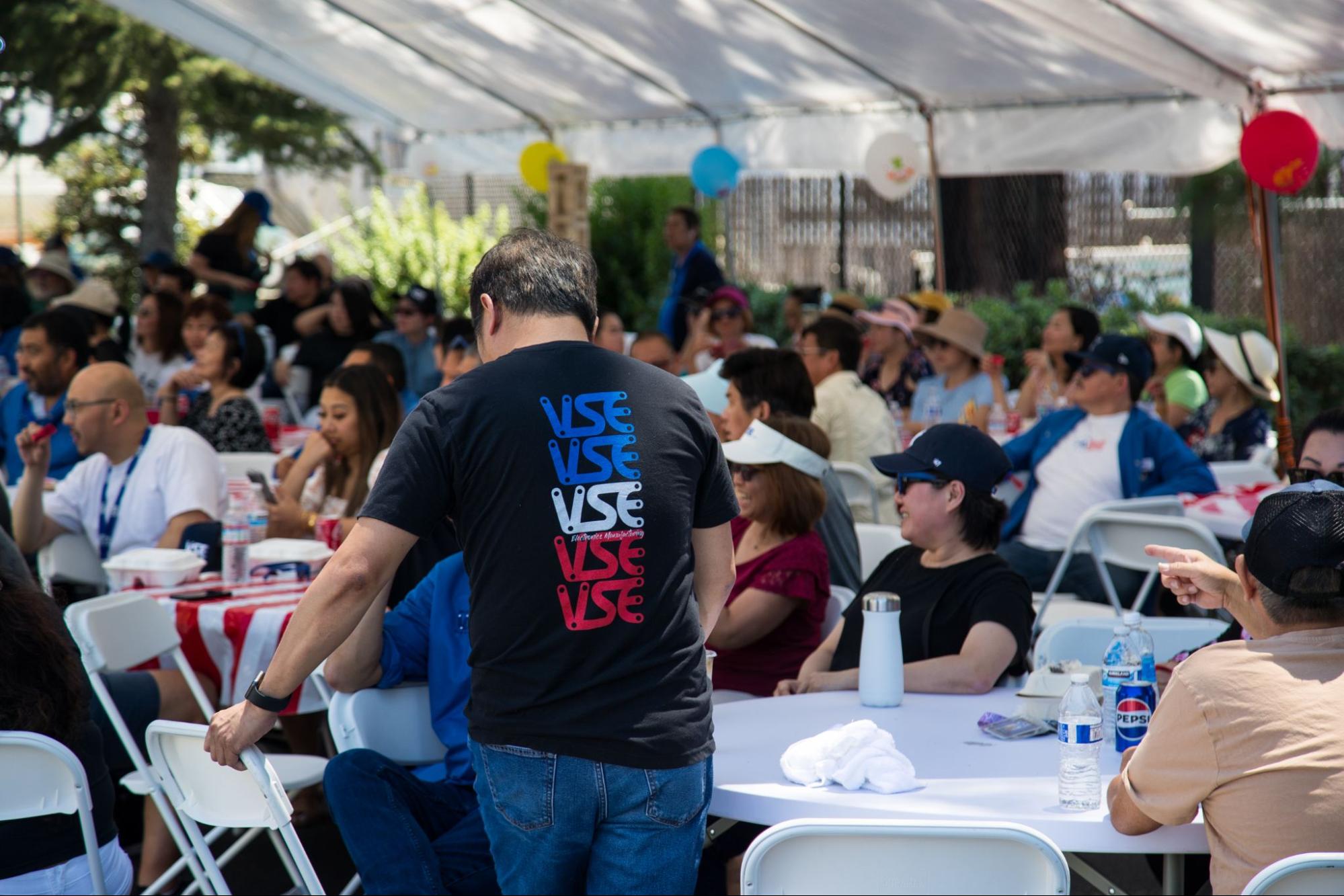 A man in a black t-shirt with a red, white, and blue "VSE" logo stands at an outdoor event. A man in a black t-shirt with a red, white, and blue "VSE" logo stands at an outdoor event.