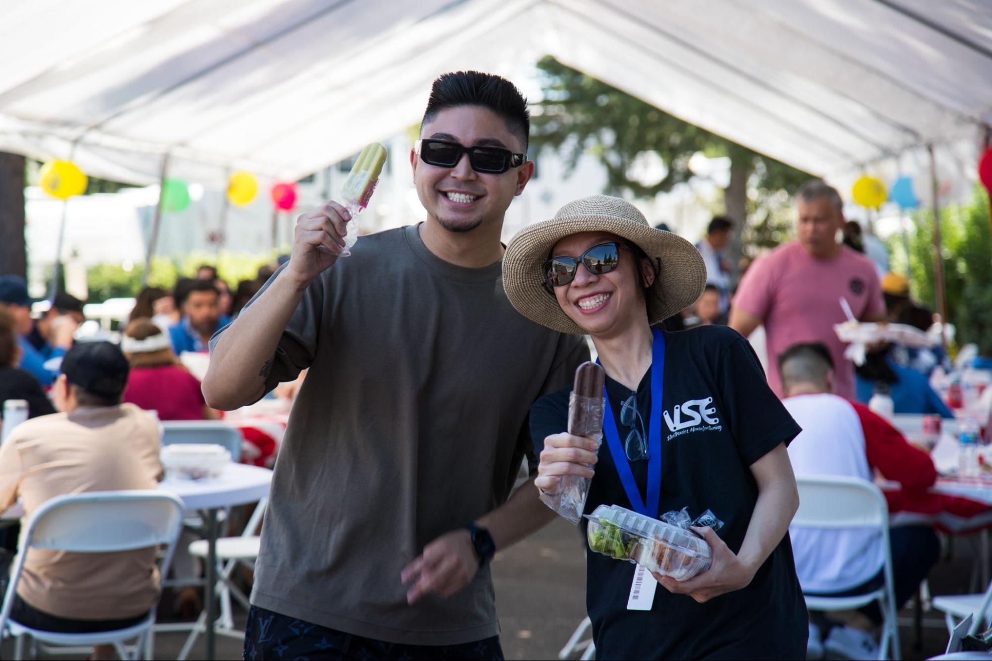 A man and a woman are smiling and holding up popsicles at an outdoor event under a tent. A man and a woman are smiling and holding up popsicles at an outdoor event under a tent.